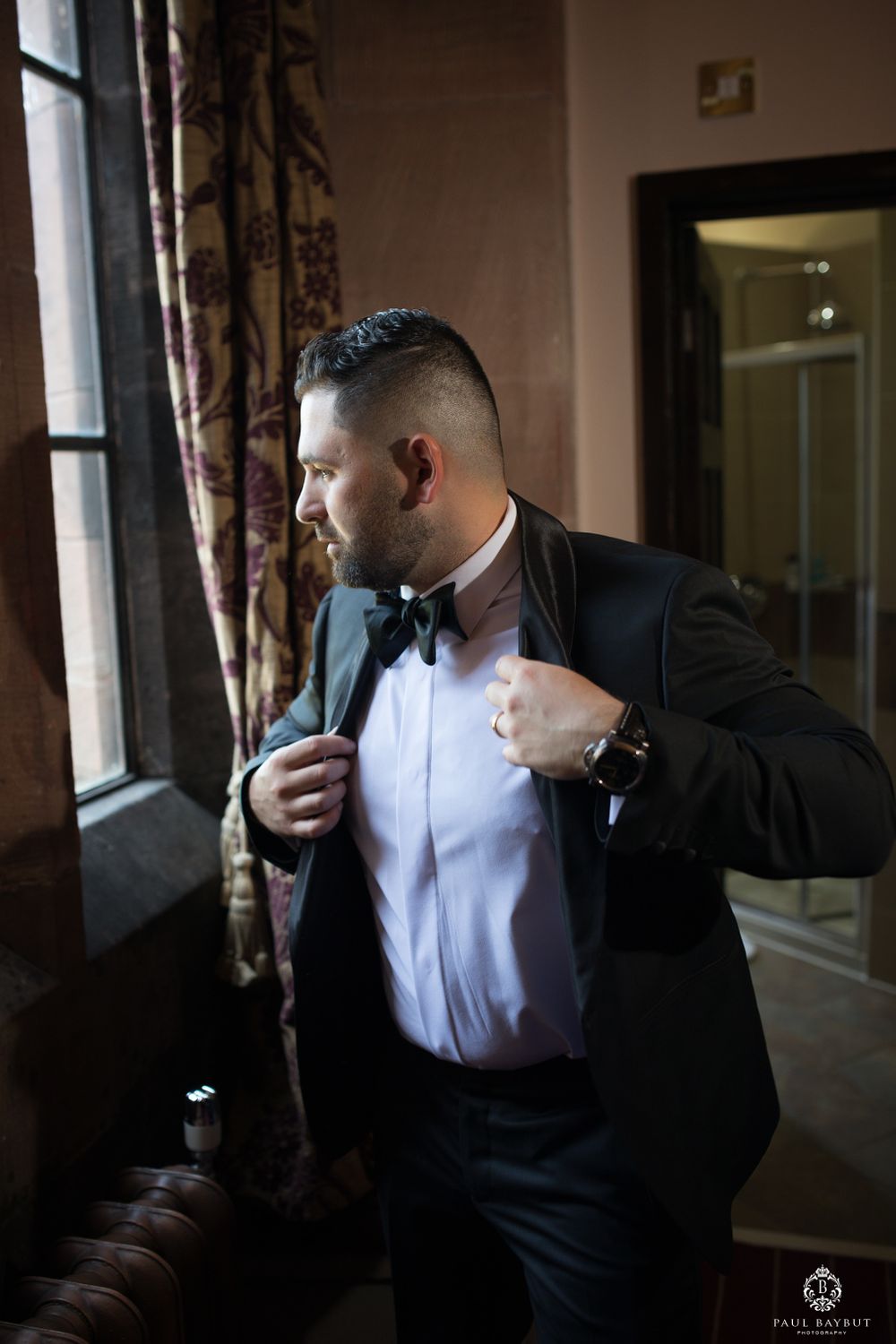 A groom puts on his formal tuxedo jacket near a window with elegant curtains.