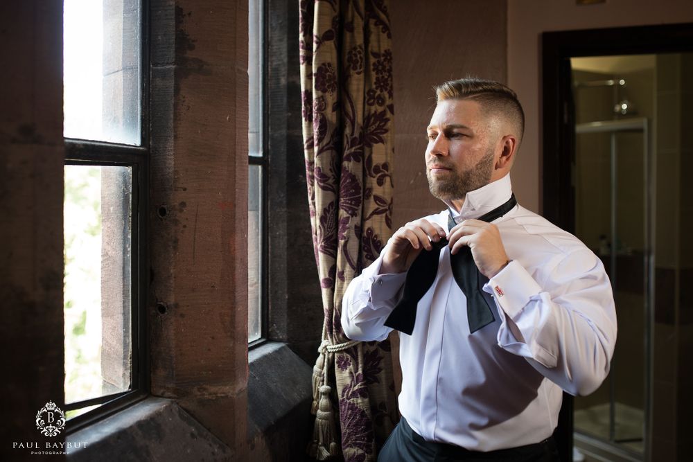 A person adjusts their bowtie while getting ready by a window in formal attire.