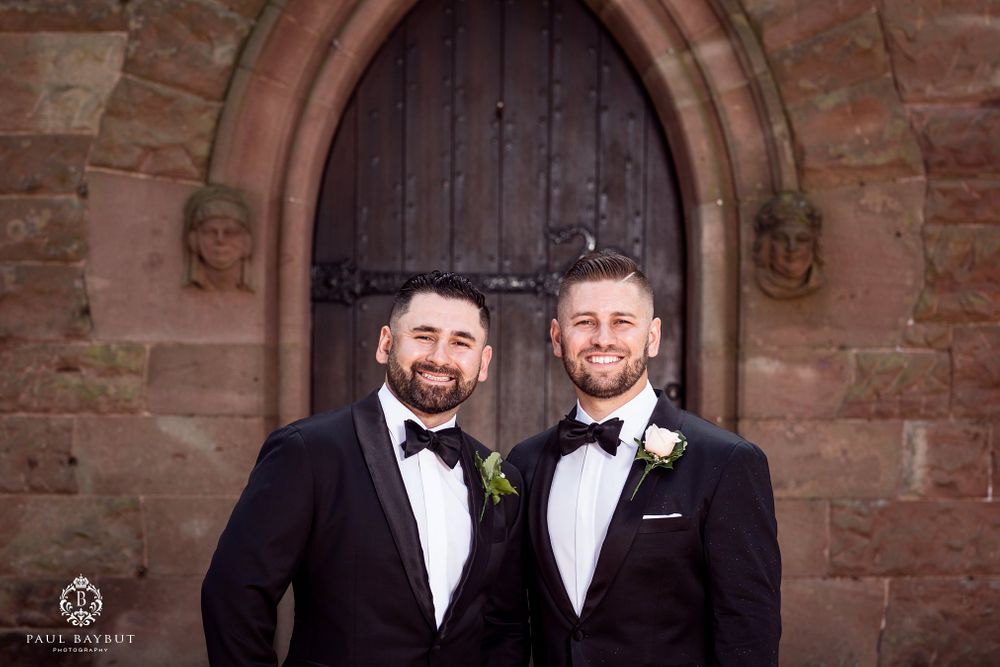 Two people in tuxedos smiling in front of an arched church doorway.