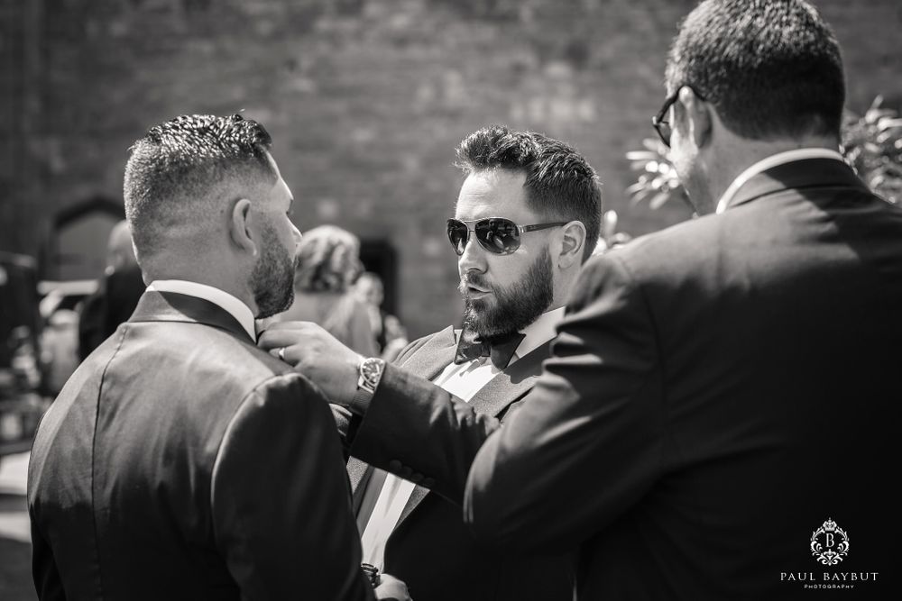 Men in formal attire and sunglasses having a candid conversation at an outdoor wedding celebration.