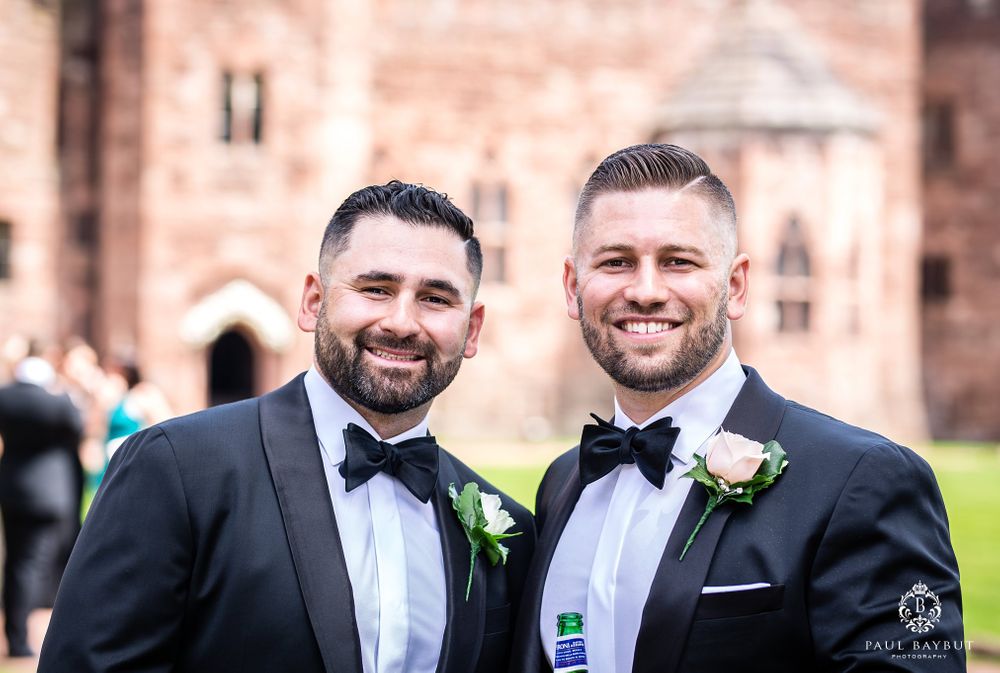 Two men in black tuxedos and bowties smiling at outdoor wedding venue.
