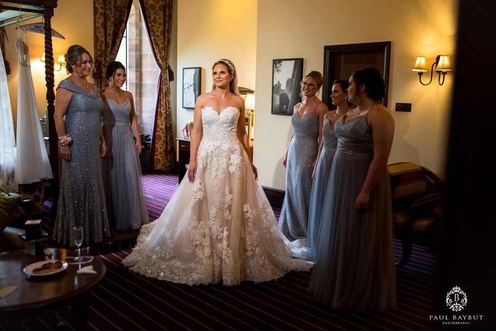 A bride in a lace wedding gown stands surrounded by bridesmaids wearing blue-gray dresses in an elegant room.