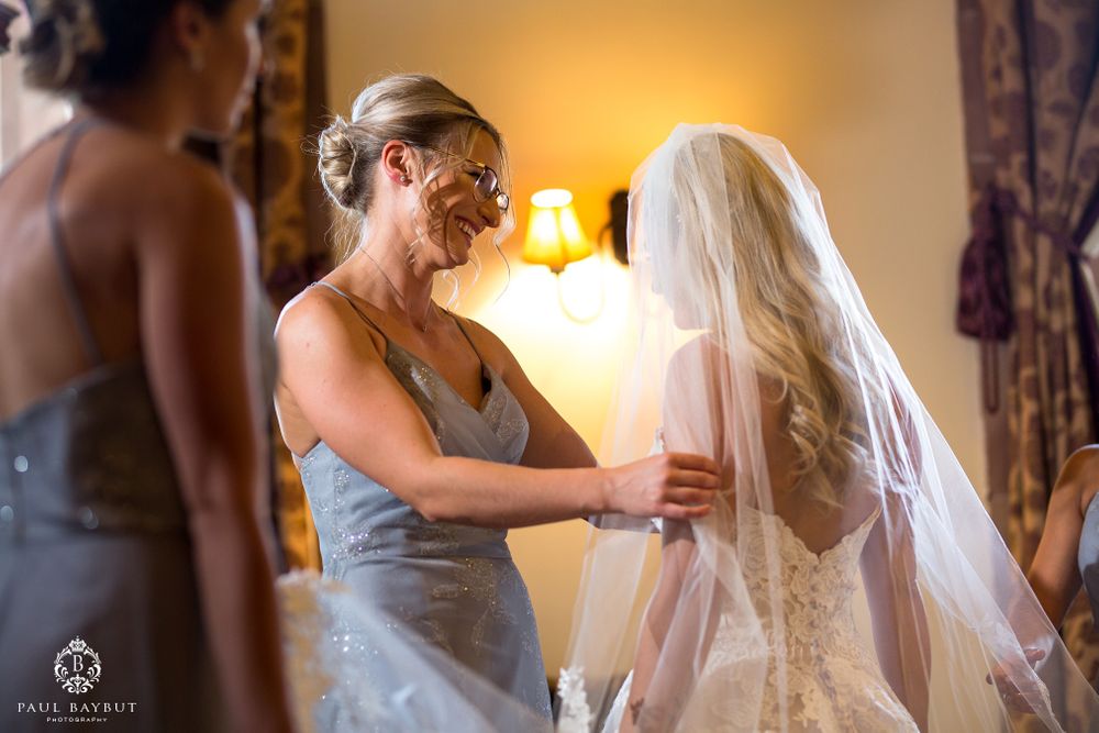 Bridesmaids in dusty blue dresses help adjust the bride's veil in a warmly lit room.