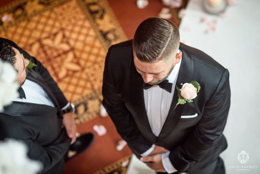 Groom in classic black tuxedo and white boutonniere photographed from above on ornate carpet.