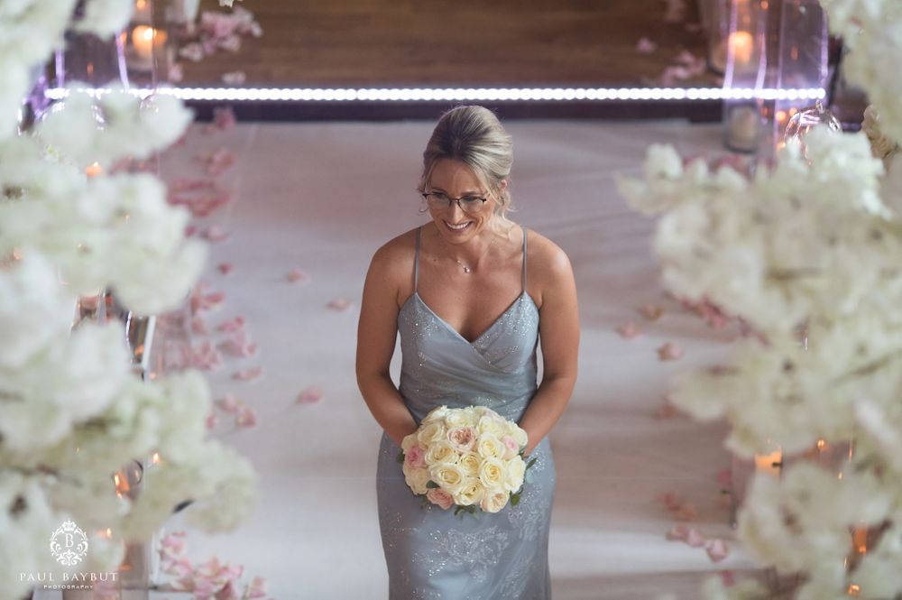 Bridesmaid in silver dress holds white bouquet surrounded by floral decorations.