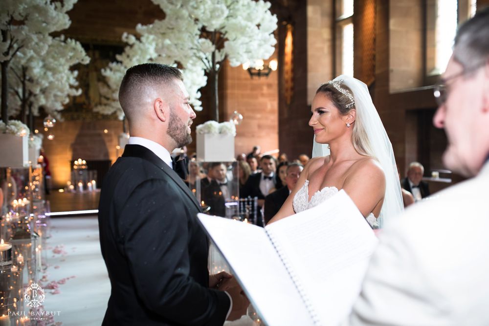 A bride and groom exchange wedding vows under elegant white floral decorations in a grand indoor venue.