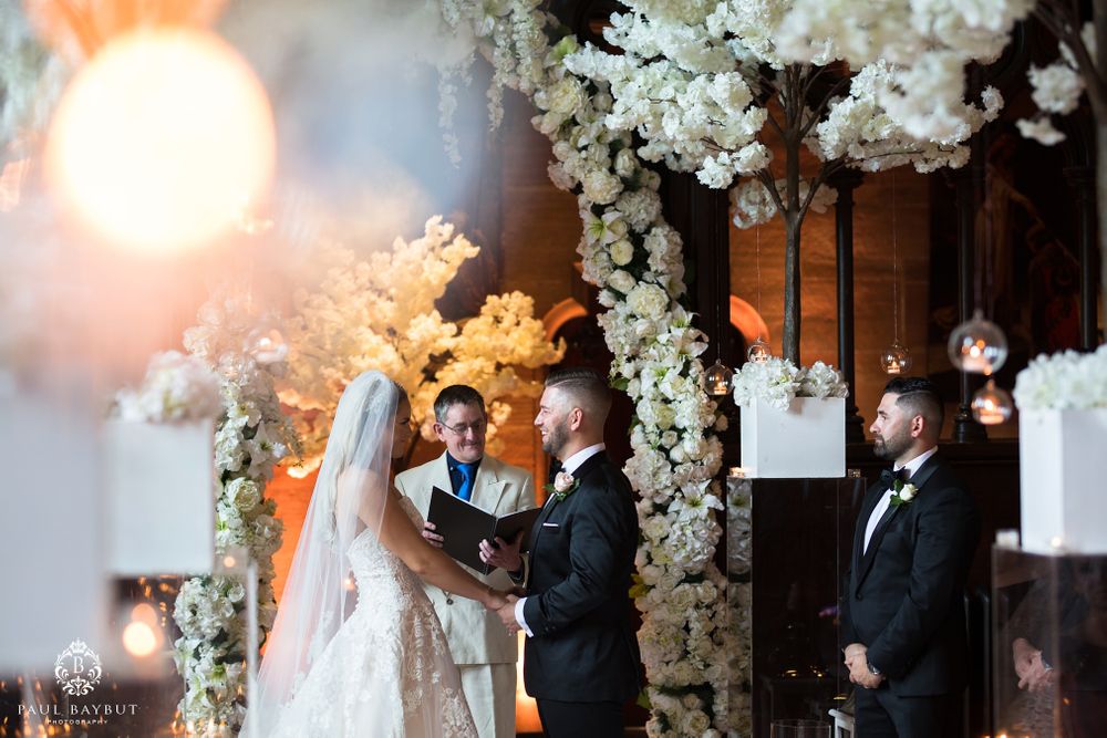 A wedding ceremony takes place beneath an elegant arch of white flowers with warm lighting.