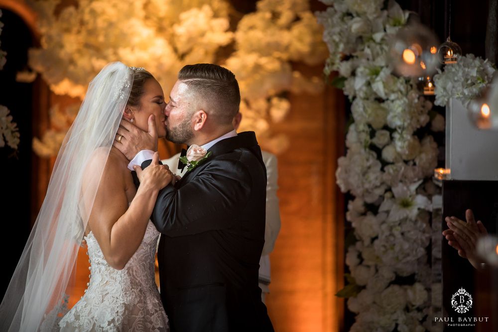 Bride and groom share romantic kiss during wedding ceremony under floral arch with warm lighting.