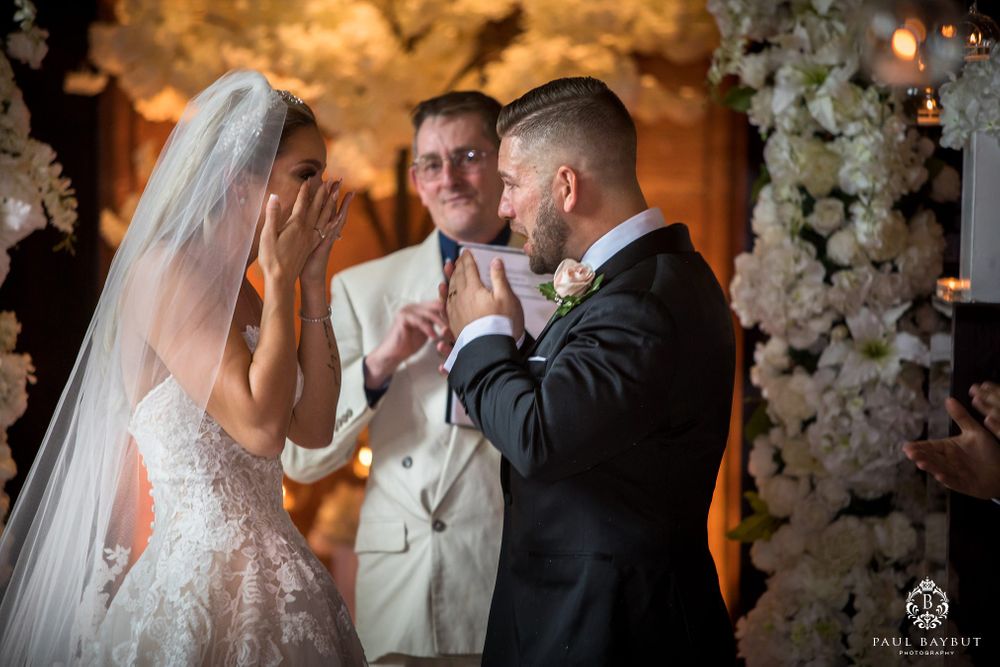 Emotional wedding ceremony moment with bride and groom exchanging vows under white floral arch.