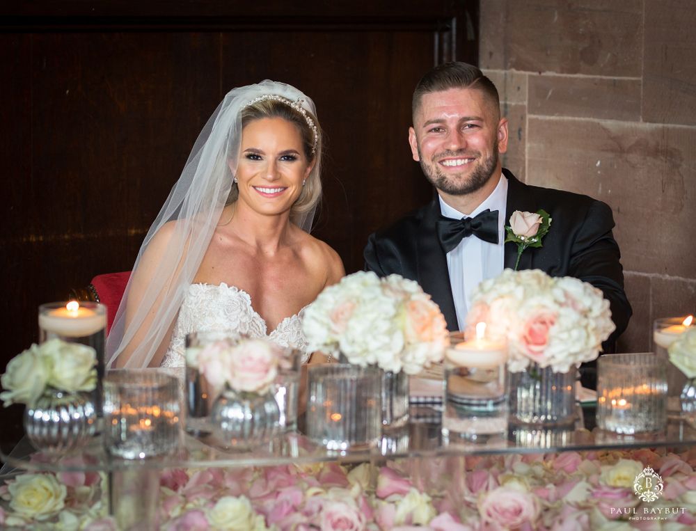 Newlyweds sit at decorated reception table with candles and floral arrangements.