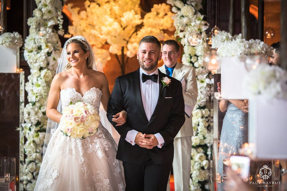 Bride and groom walk down aisle under elaborate white floral arch with warm lighting.