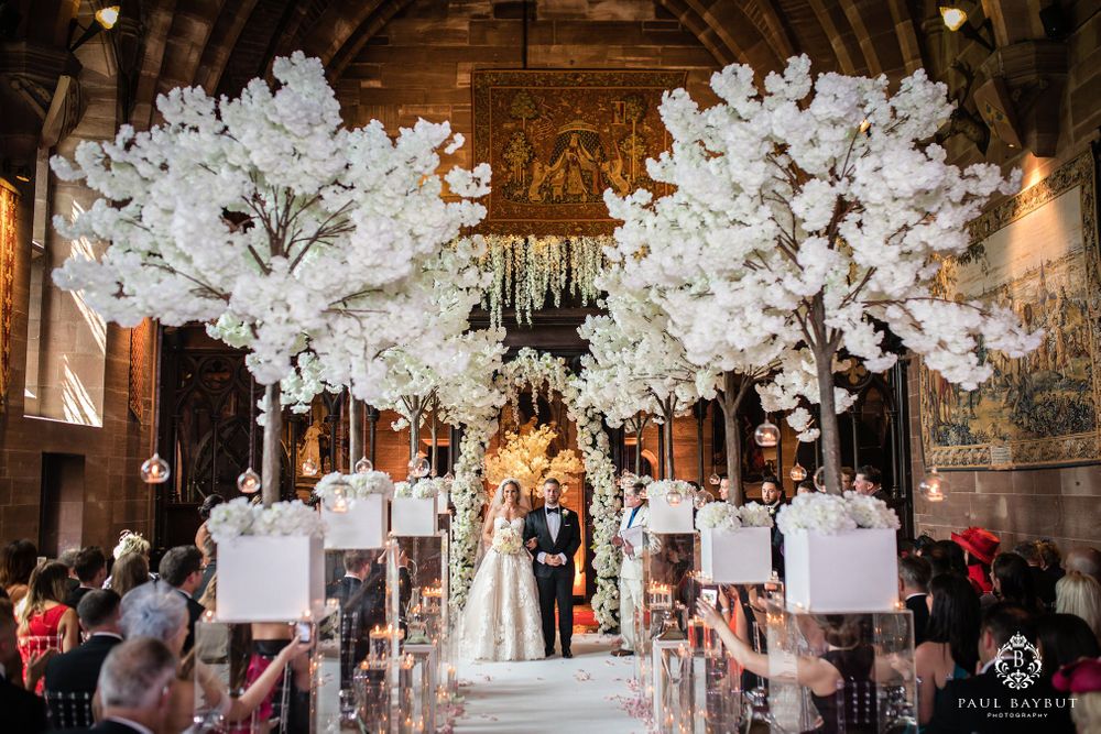 Elegant wedding ceremony aisle with tall white cherry blossom trees and hanging lanterns in rustic wooden interior.