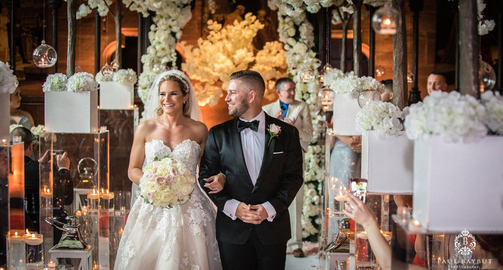 Wedding ceremony under ornate white floral arch with romantic lighting in rustic venue.