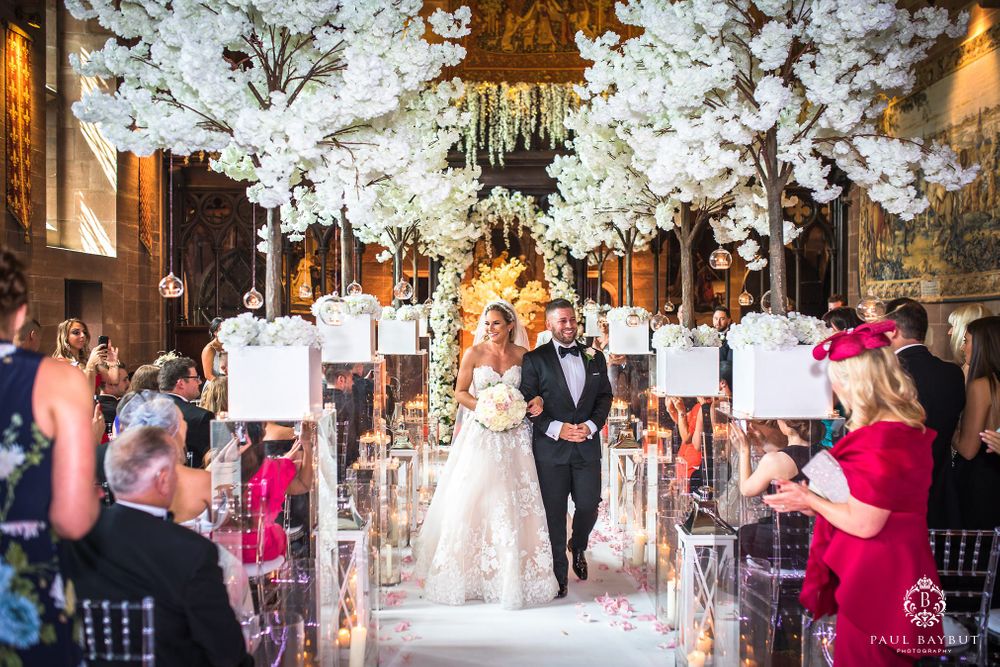 Wedding couple walks down aisle decorated with white cherry blossoms as guests capture the moment.