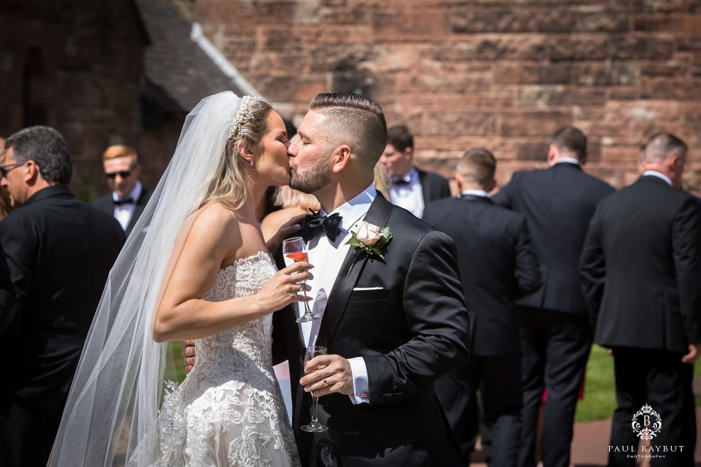 A romantic wedding kiss shared in front of a historic brick wall with guests gathered in the background.