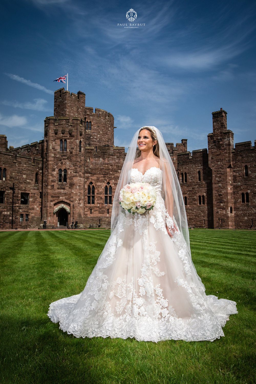 A bride in a lace wedding gown stands on castle grounds holding a white bouquet against blue skies.