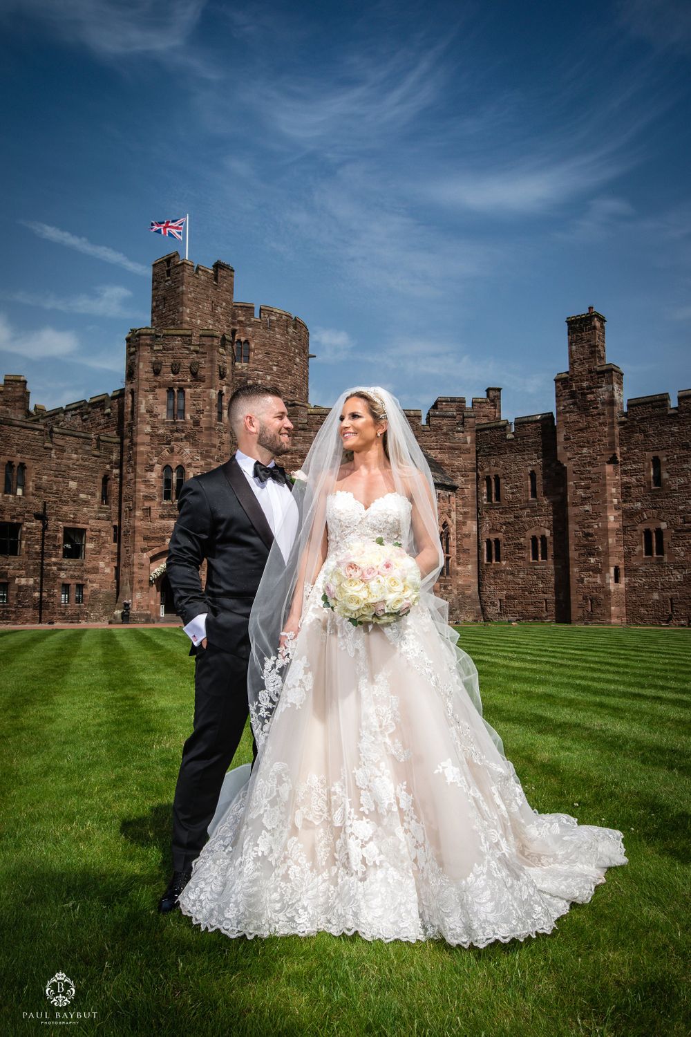 A wedding couple poses in front of a majestic castle with a British flag flying overhead on a sunny day.