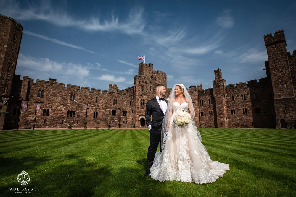 A wedding couple poses on the manicured lawn of a medieval castle under bright blue skies.