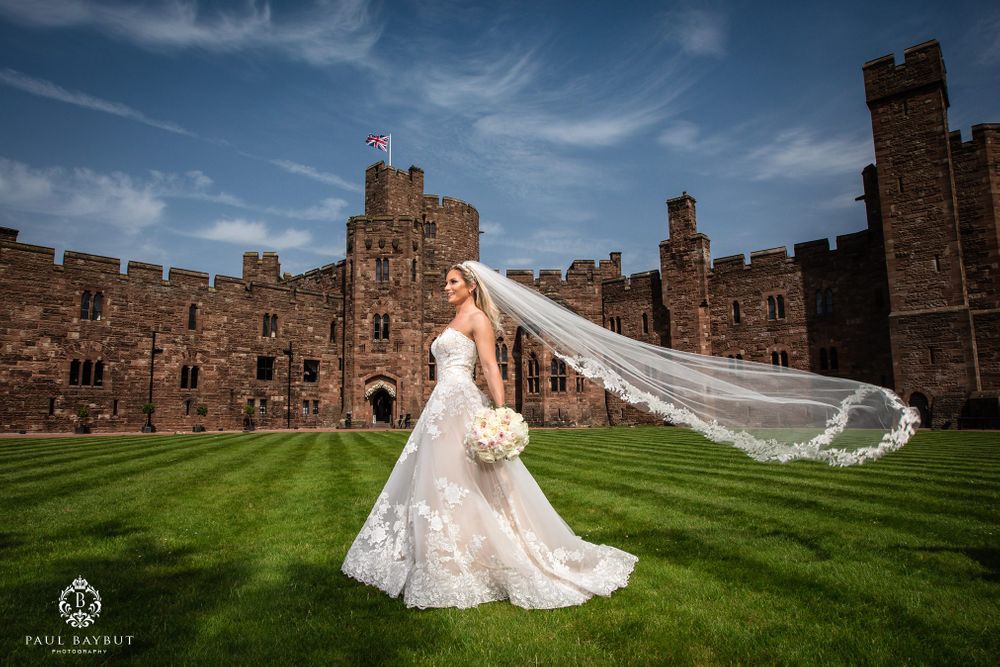 A bride with flowing veil stands on castle grounds as her dress and veil catch the wind dramatically.