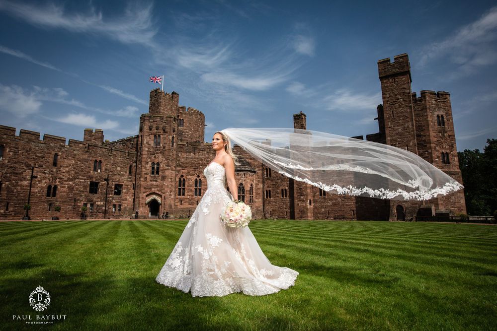 A bride's long veil flows in the wind against the backdrop of a historic castle with stone walls.