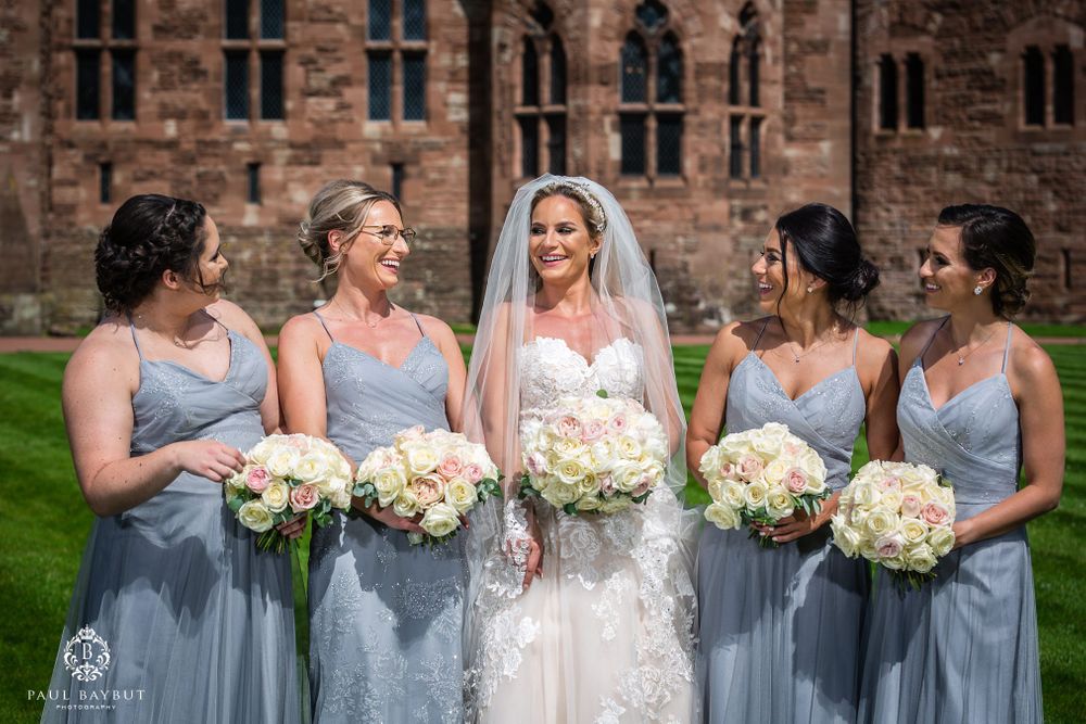 Wedding party with bridesmaids in light blue dresses holding white flower bouquets by castle walls.
