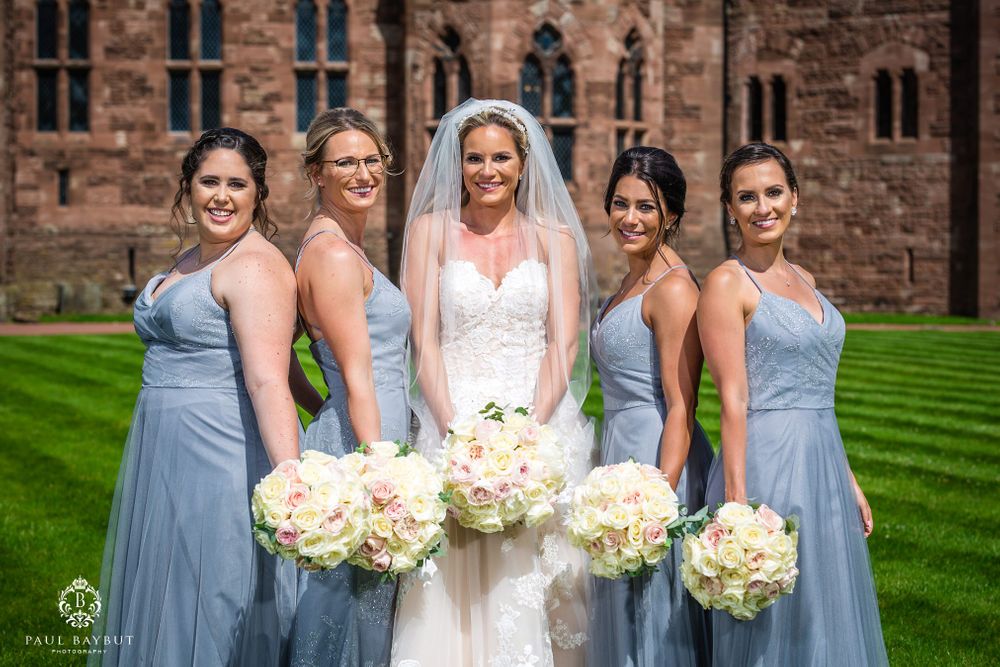 Bridesmaids in flowing blue dresses pose with bouquets against gothic castle architecture.