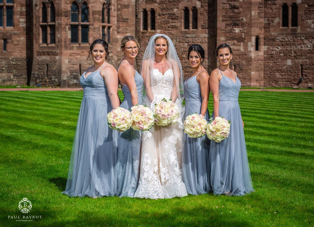 Wedding party in blue bridesmaid dresses poses with bouquets against historic castle backdrop.