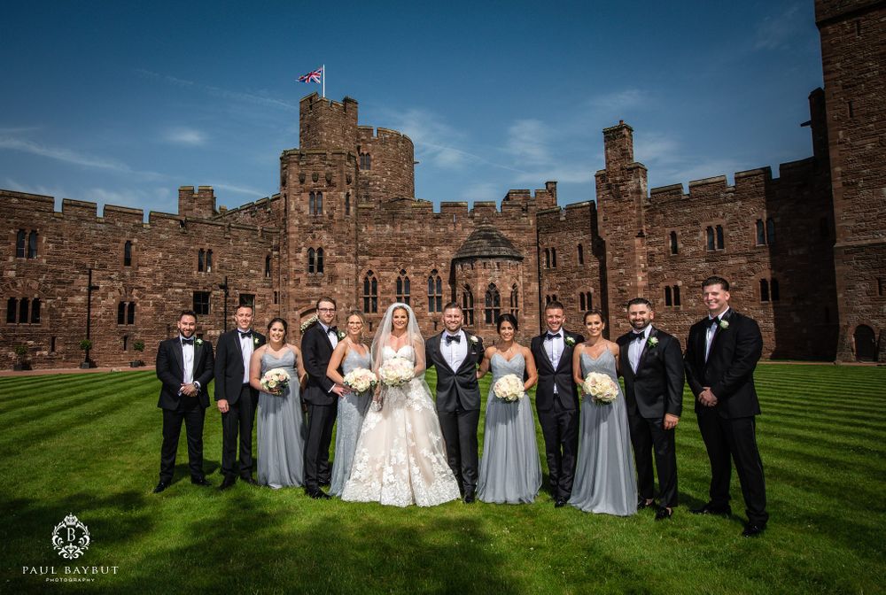 Wedding party in formal attire gathers on castle lawn with stone fortress architecture behind them.