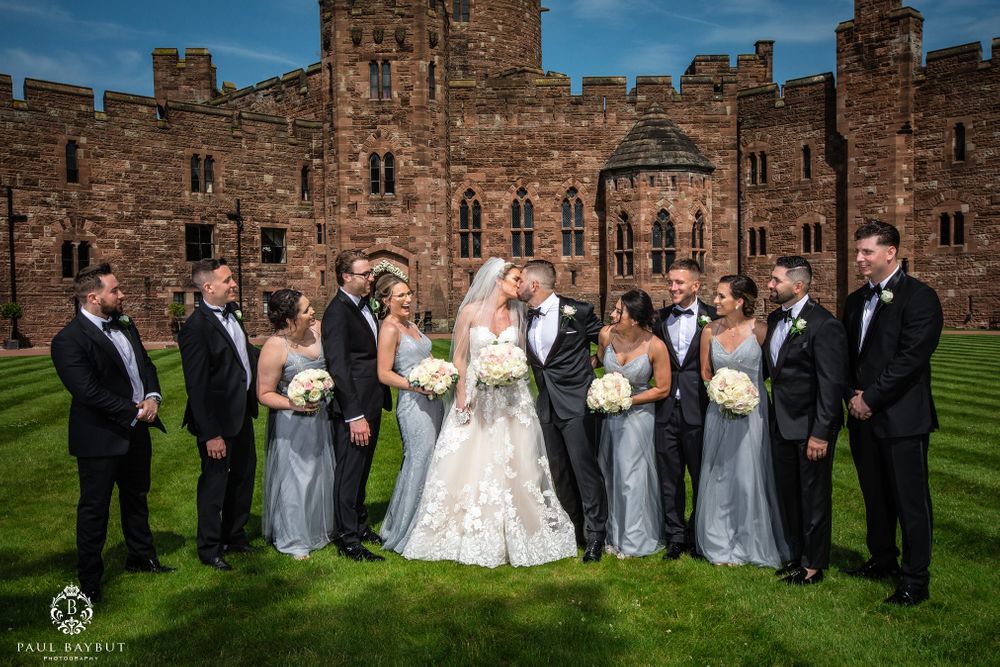 Large wedding party poses for formal photo in front of medieval castle architecture.