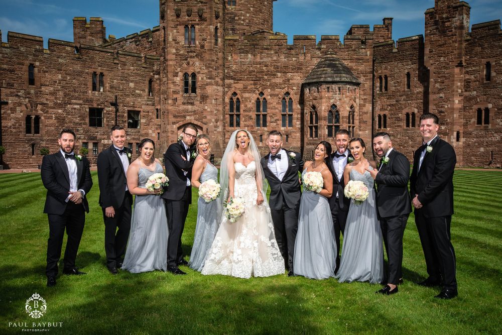 Wedding group portrait with bridesmaids in gray and groomsmen in black against castle walls.