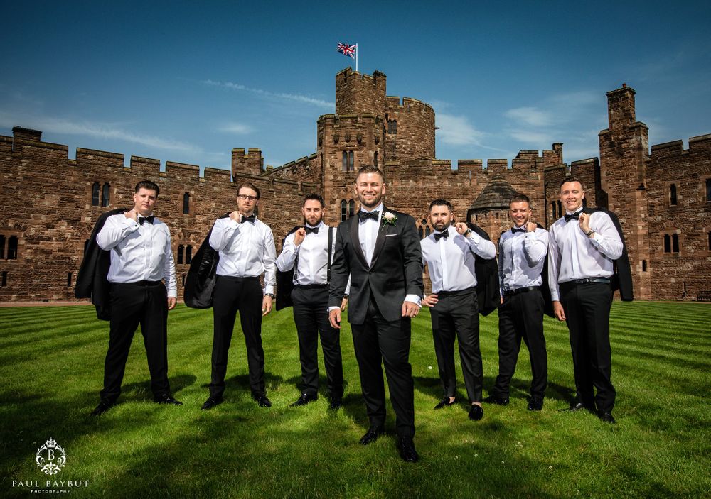 Groomsmen in white shirts and black attire pose against castle ramparts under blue sky.