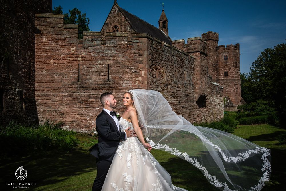Wedding couple poses as veil flows dramatically against backdrop of historic castle.