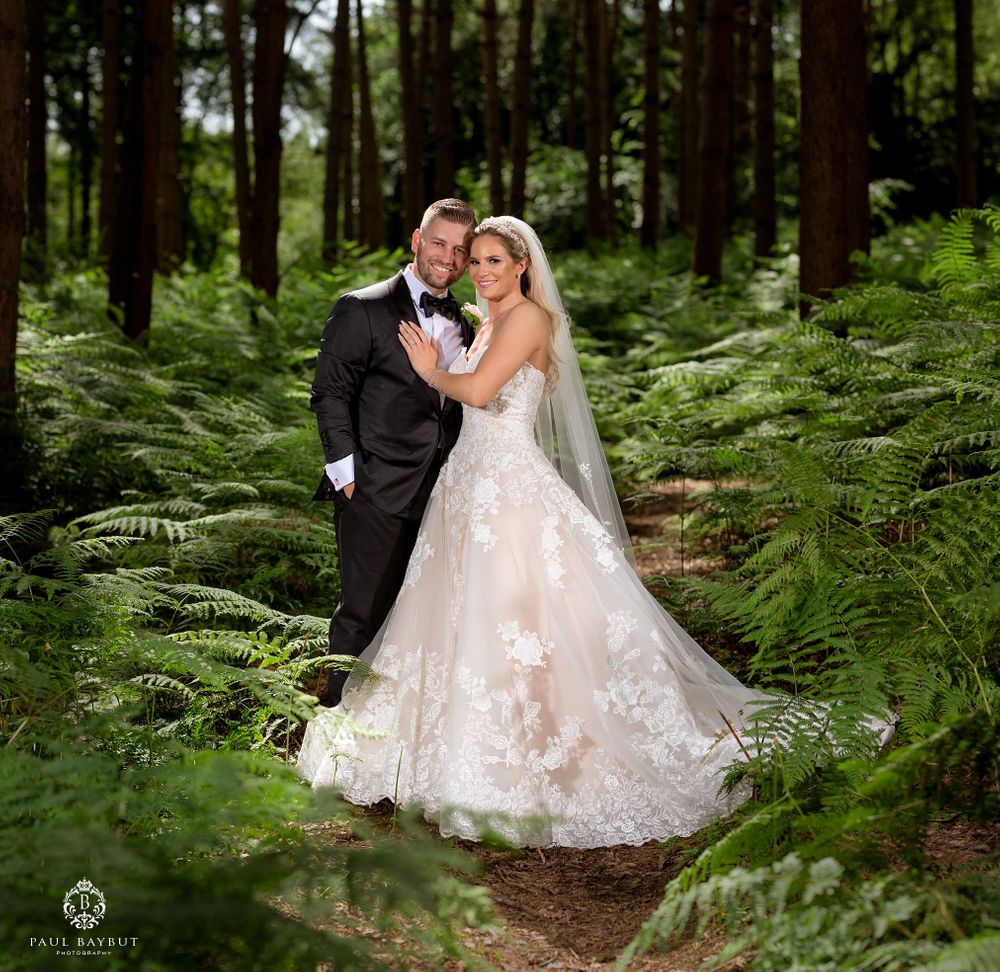 A wedding couple poses on a forest path surrounded by lush green ferns and tall trees.