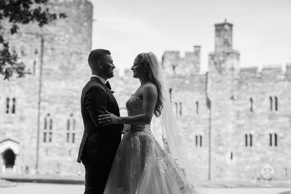 A wedding couple embraces in front of a majestic stone castle with historic architecture.