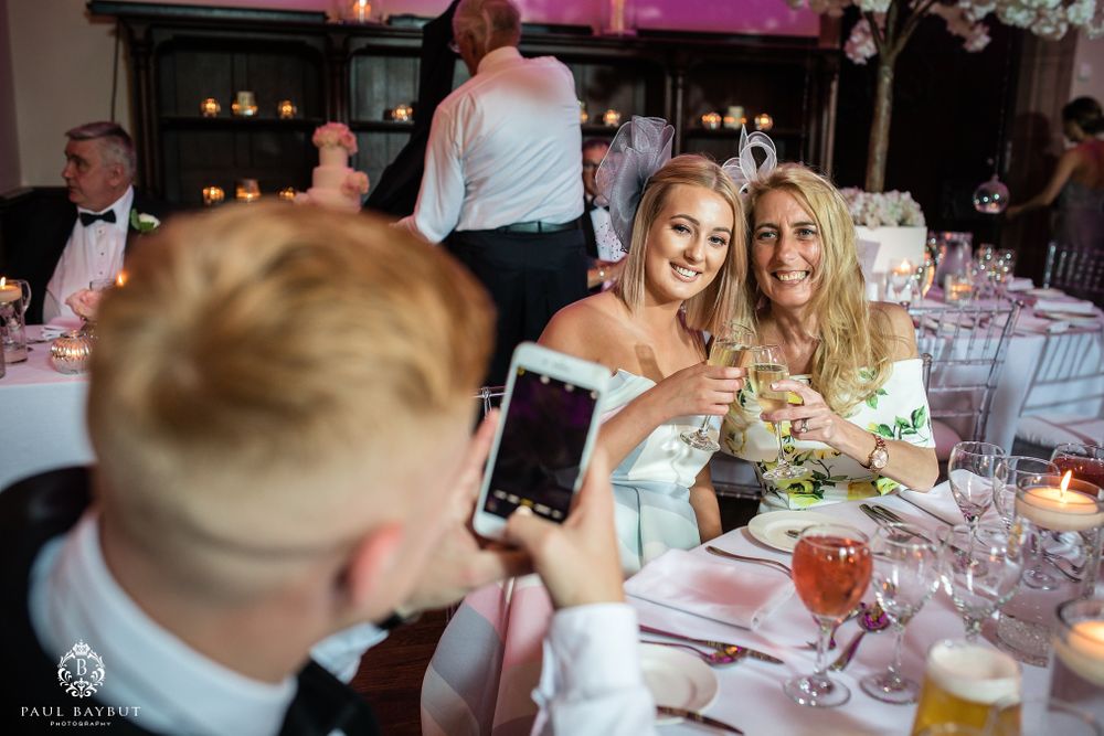 Wedding reception guests seated at decorated tables taking photos with smartphones.