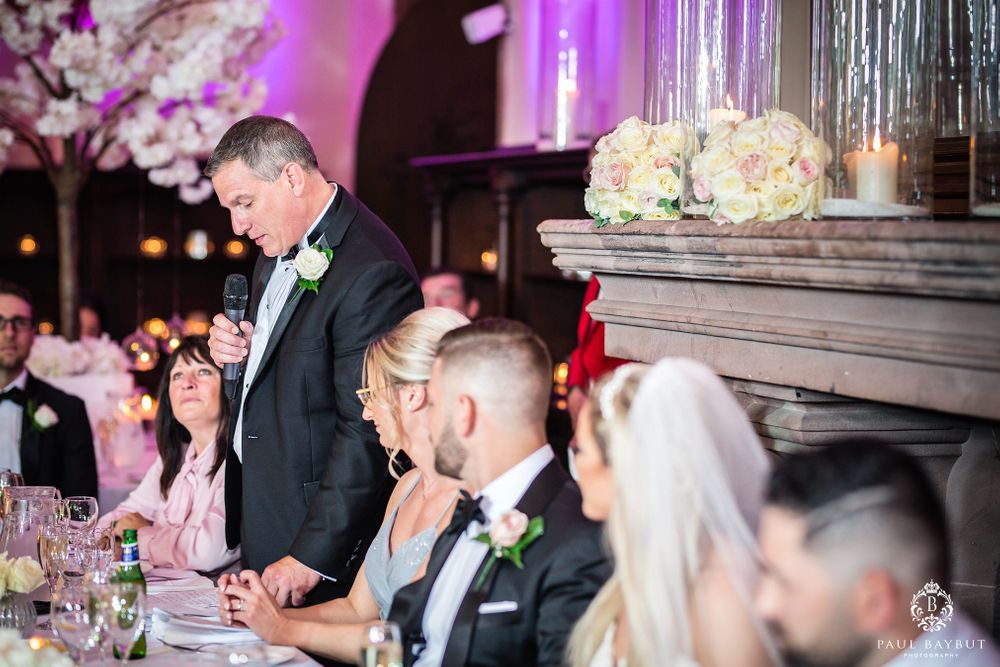 Wedding guests gather at an elegantly decorated reception table with pink uplighting and floral centerpieces.