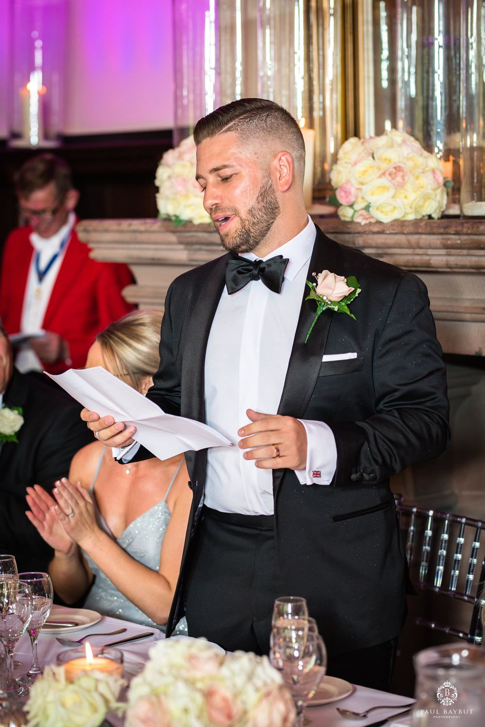 A formal speech being delivered at an elegant wedding reception with pink uplighting and floral decor.