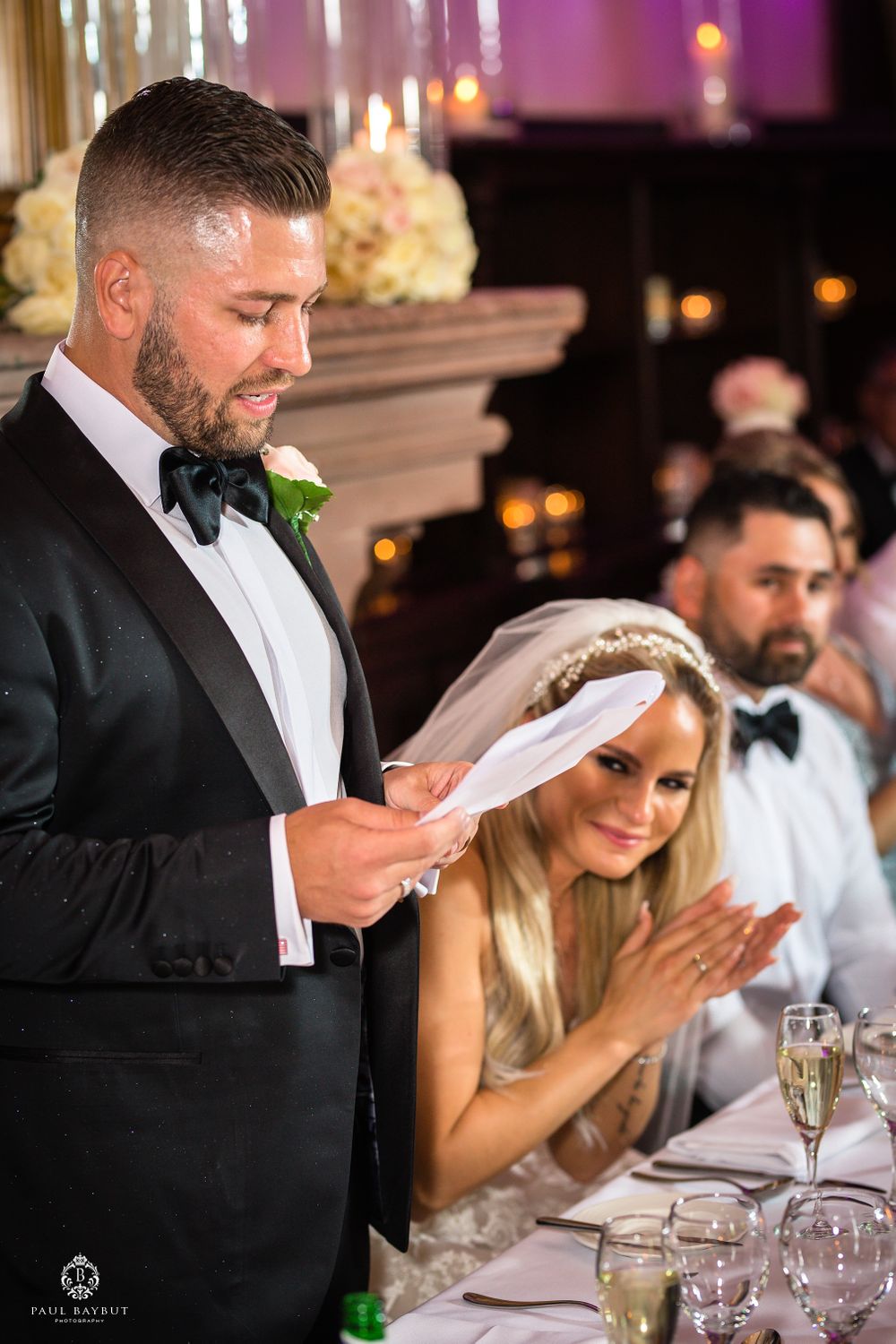 A groom delivers a wedding speech at the reception while seated guests listen at an elegantly decorated venue.
