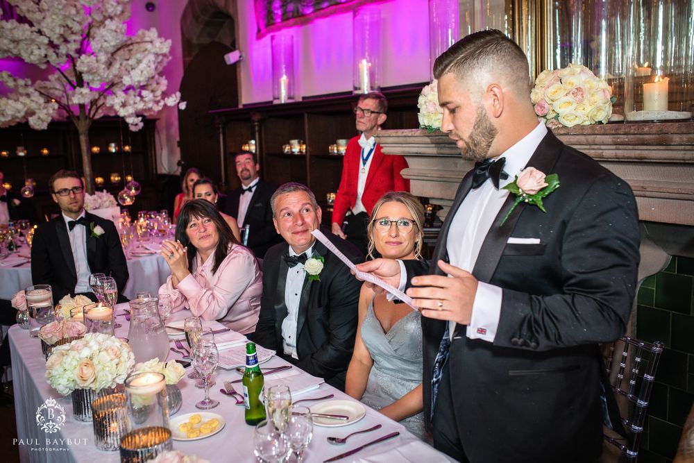 Wedding guests react during a speech at an elegant reception with pink uplighting and floral centerpieces.