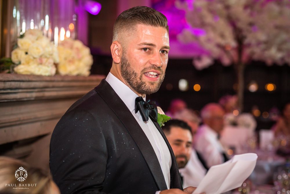 A speaker in a black tuxedo with a boutonniere delivers remarks at an elegant evening reception.