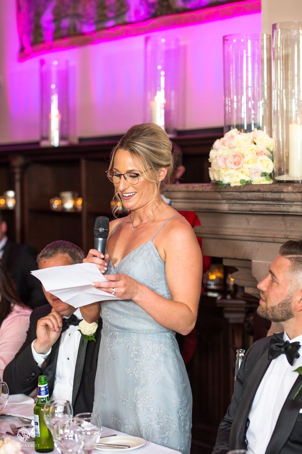 Bridesmaid in pale blue dress gives speech at elegant wedding reception with pink accent lighting.