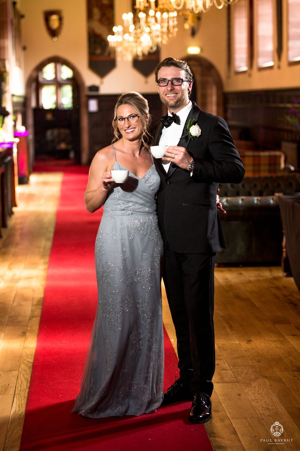 Formal couple posing with drinks in an elegant hallway with chandeliers.