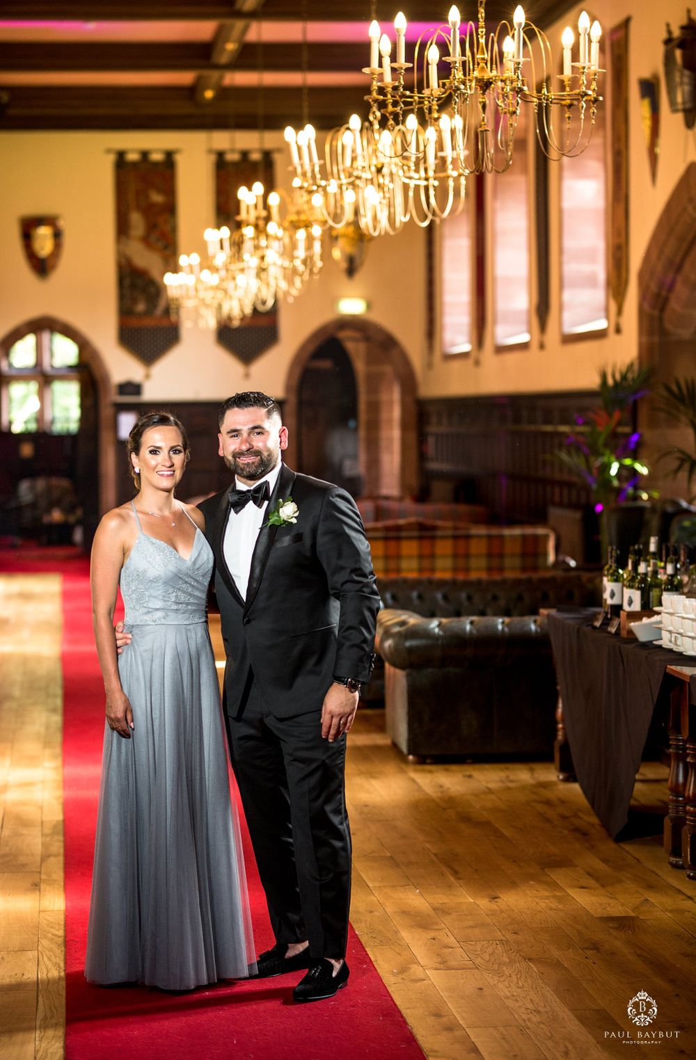 Elegant couple standing in formal attire under ornate chandeliers in a grand hallway.