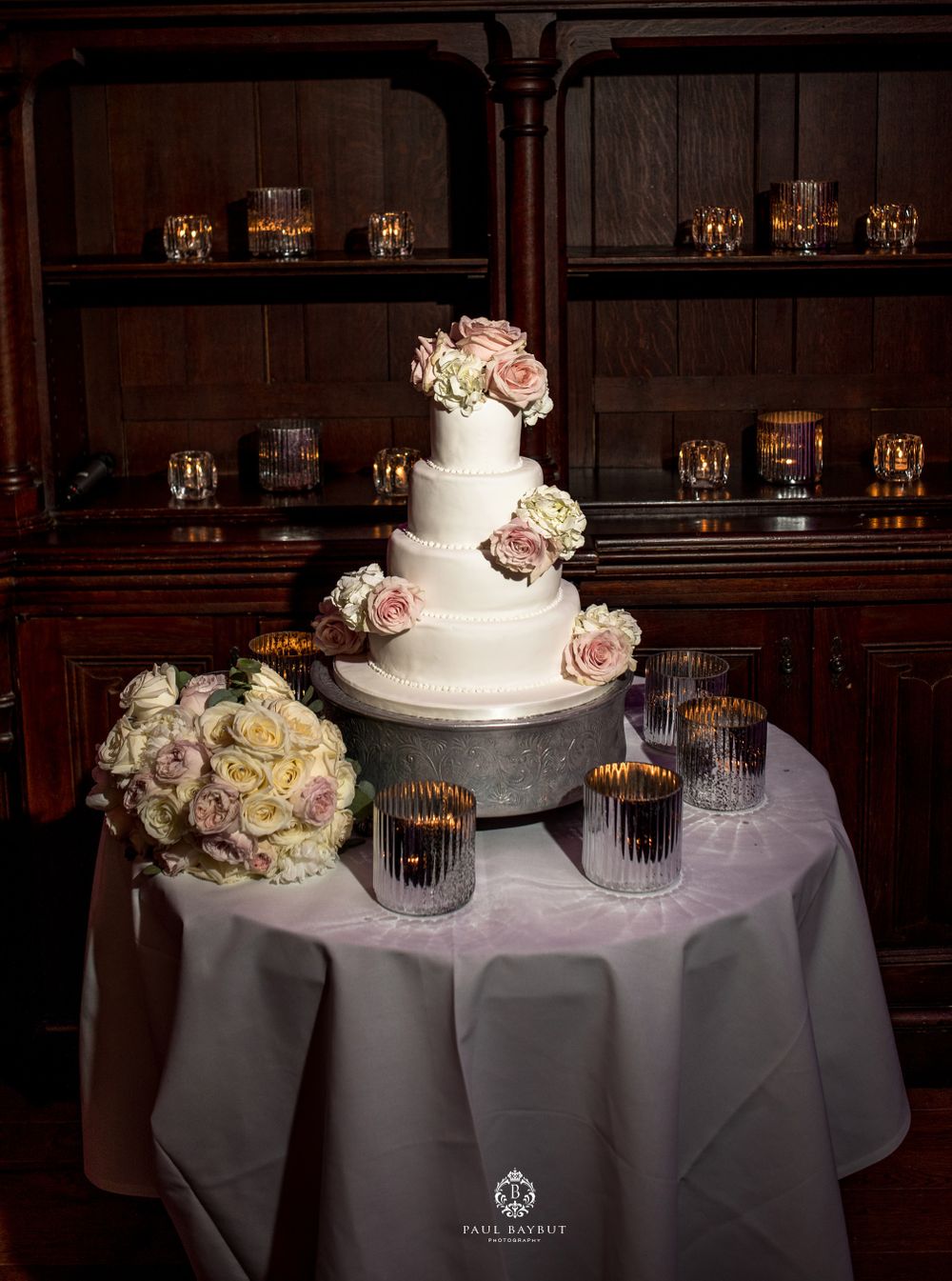 Elegant white wedding cake with pink roses displayed on table with candles and wooden shelving in background.