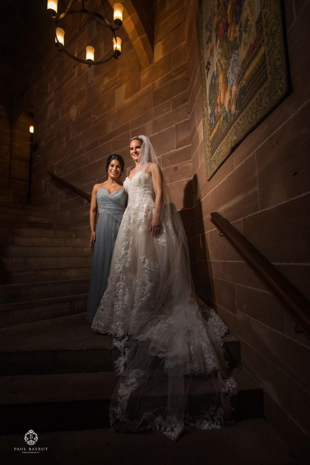 A bride in lace gown and bridesmaid in gray dress pose on stone stairs with dramatic lighting.
