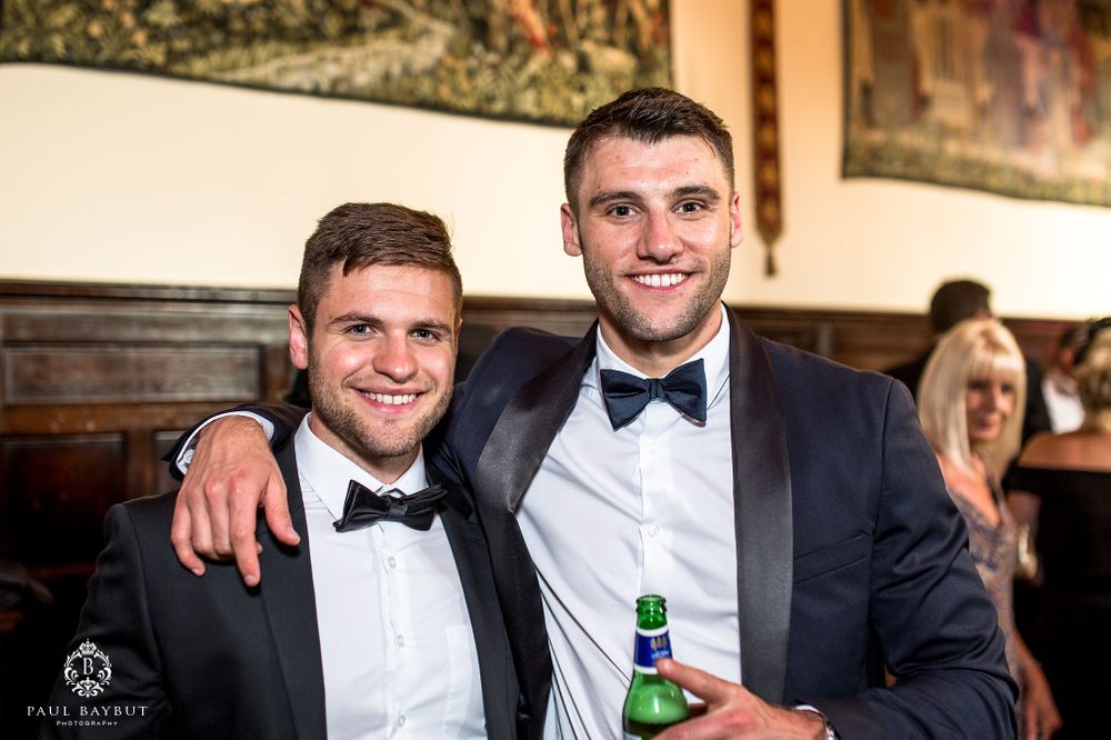 Two guests in black tuxedos and bow ties pose together at an elegant wedding reception.