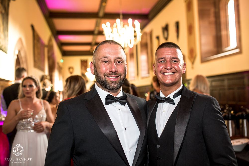 Two guests in tuxedos share a joyful moment at an elegant indoor wedding reception.