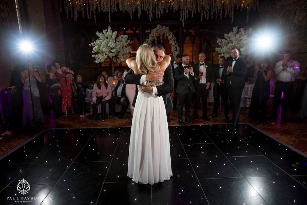 Newlyweds share their first dance on an illuminated black dance floor under ornate white floral arrangements.
