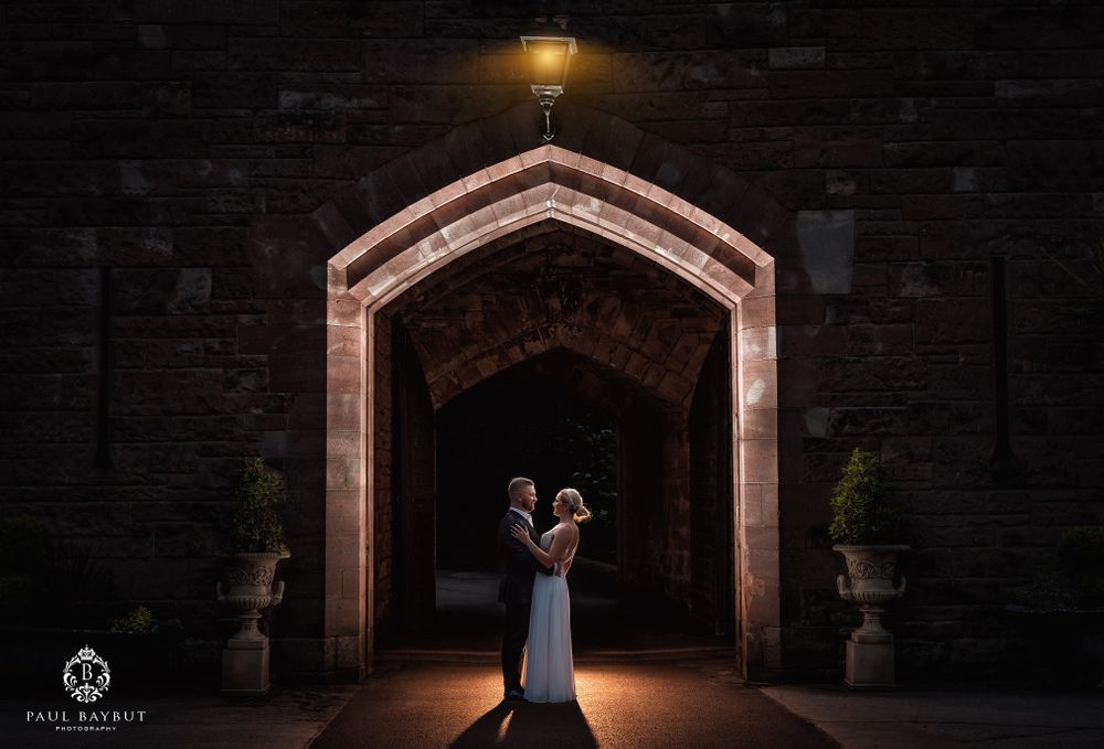 Romantic couple stands in archway of historic stone building illuminated by warm light.