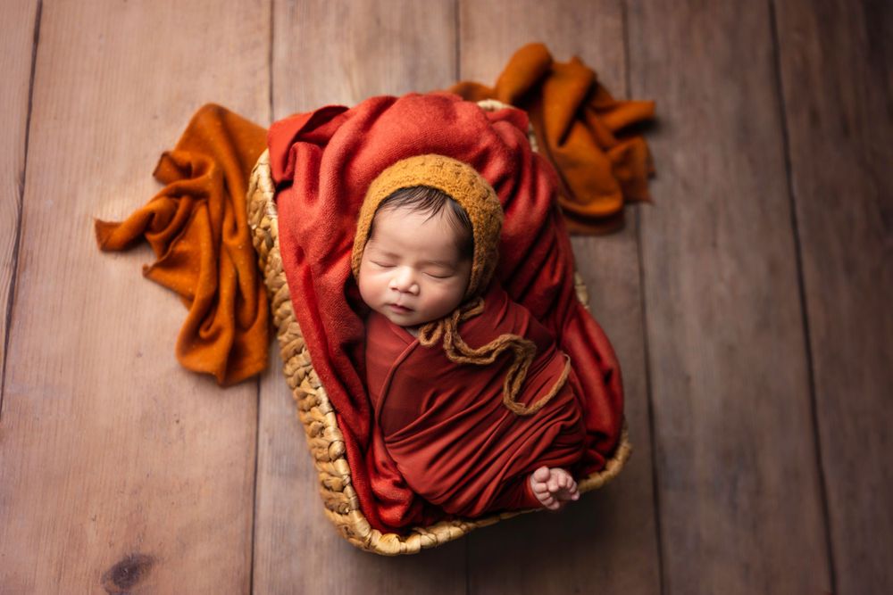 A sleeping newborn wrapped in red and orange blankets rests on wooden flooring.